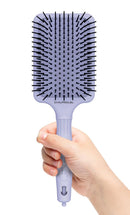 Lavender paddle hair brush held in hand, showing black bristles and cushioned base